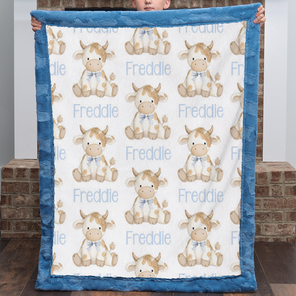 A baby blanket held up by a child, featuring a white background with a repeating pattern of cute cartoon cows and the name 'Freddie' in light blue text, finished with a thick blue plush border.