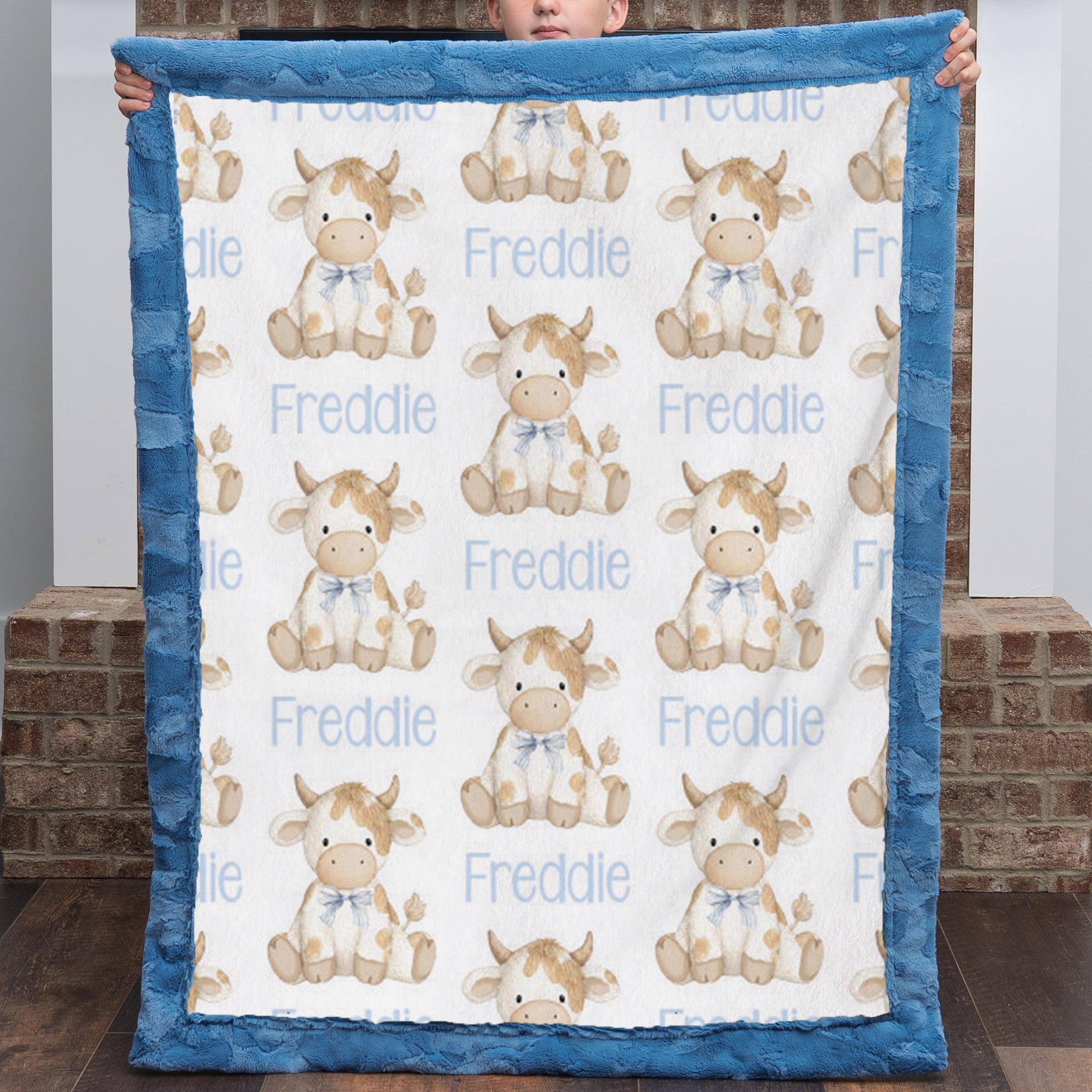 A baby blanket held up by a child, featuring a white background with a repeating pattern of cute cartoon cows and the name 'Freddie' in light blue text, finished with a thick blue plush border.