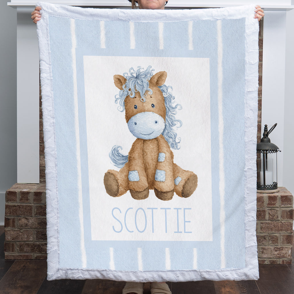 A person holds up a personalized baby blanket featuring a cartoon illustration of a brown horse with a blue mane and the name 'SCOTTIE' printed below, set against a light blue and white striped background.