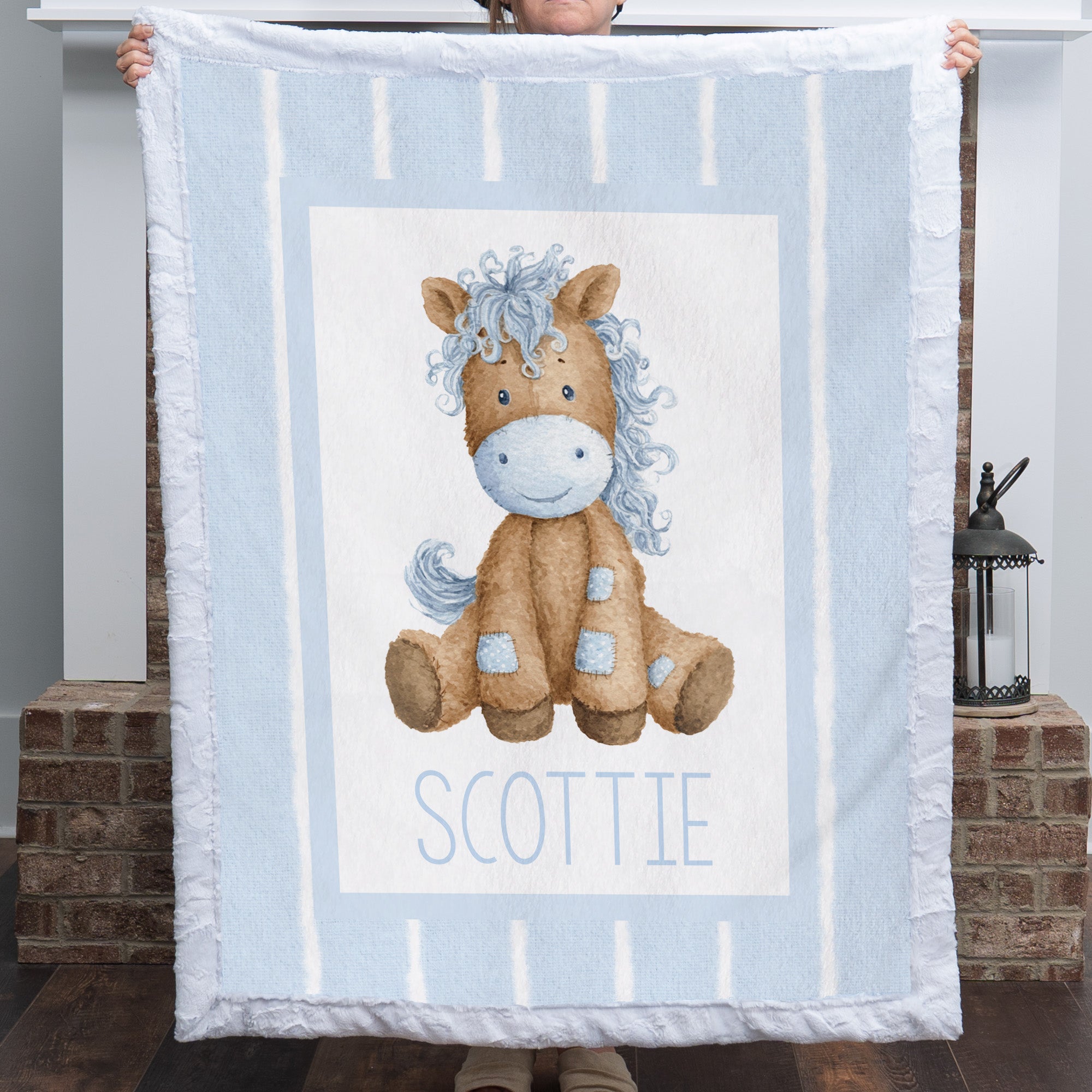 A person holds up a personalized baby blanket featuring a cartoon illustration of a brown horse with a blue mane and the name 'SCOTTIE' printed below, set against a light blue and white striped background.