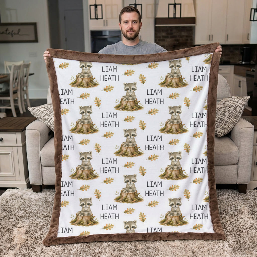 A man stands in a living room holding up a large custom fleece blanket. The blanket features a white background printed with a repeating pattern of gray-brown raccoons sitting on logs, gold leaves, and the text 'LIAM HEATH'.