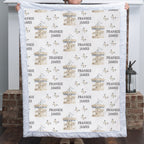 A person holds up a custom quilted blanket featuring a repeating pattern of carousel illustrations, butterflies, and the personalized name 'FRANKIE JAMES' in gray text. The blanket has a white quilted border and is shown against a home interior background with brick pillars.