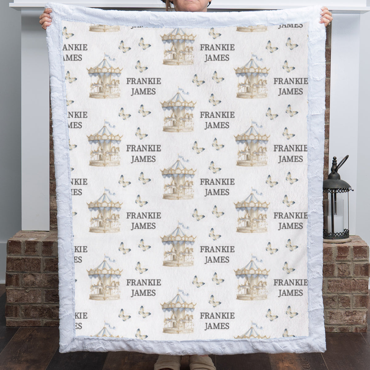 A person holds up a custom quilted blanket featuring a repeating pattern of carousel illustrations, butterflies, and the personalized name 'FRANKIE JAMES' in gray text. The blanket has a white quilted border and is shown against a home interior background with brick pillars.