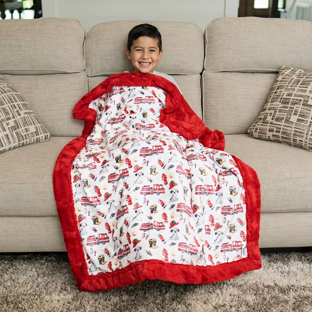 Smiling boy on sofa wrapped in firefighter Luxe Cuddle blanket with fire truck and Dalmatian print