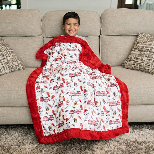 Smiling boy on sofa wrapped in firefighter Luxe Cuddle blanket with fire truck and Dalmatian print
