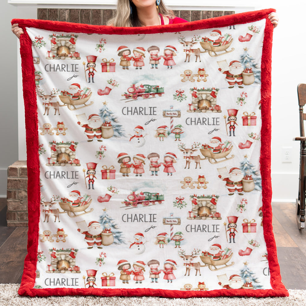 A person holds up a large personalized Christmas blanket with a red plush border and a white background printed with Santa Claus, sleighs, trees, gifts, and the name 'CHARLIE' repeated across the front.