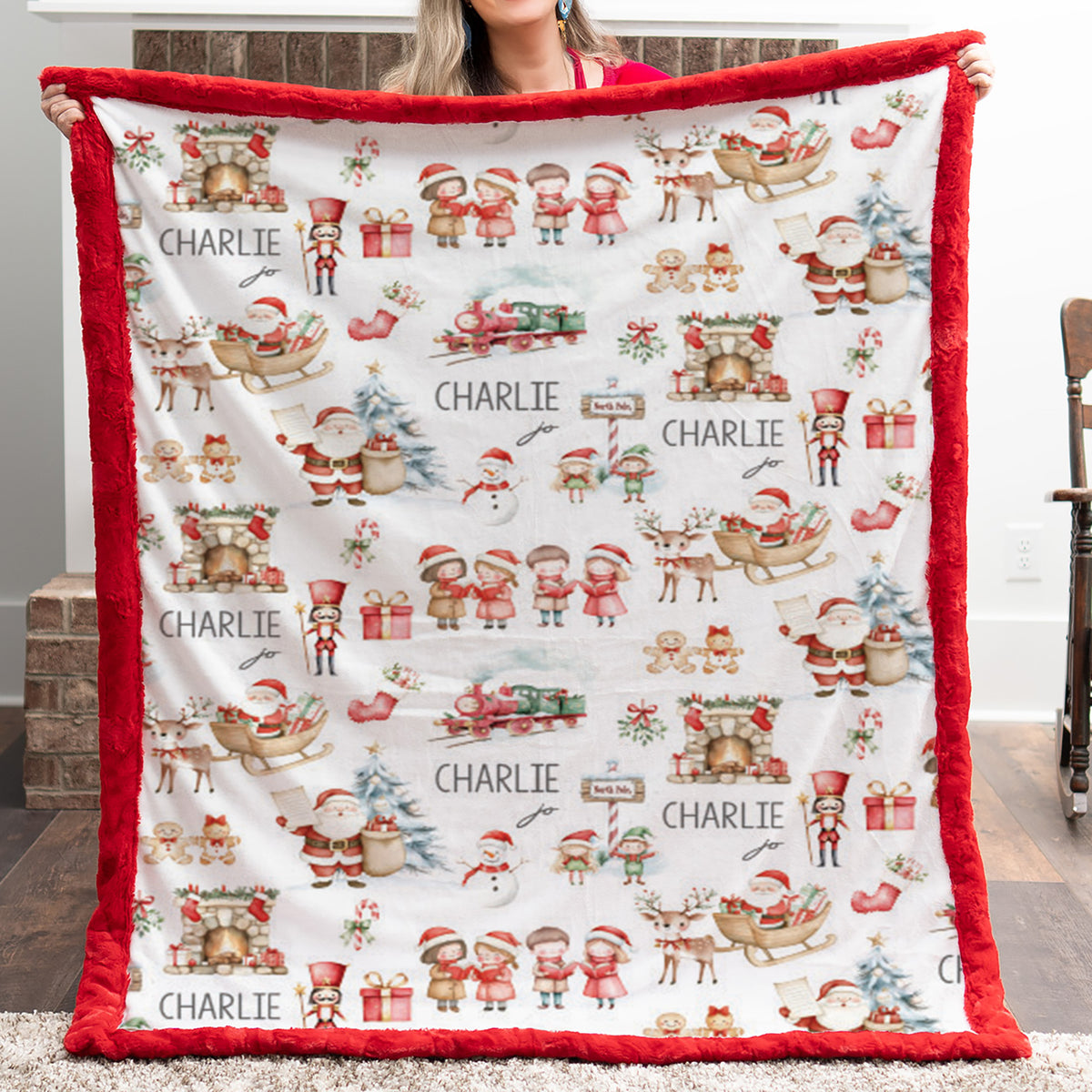 A person holds up a large personalized Christmas blanket with a red plush border and a white background printed with Santa Claus, sleighs, trees, gifts, and the name 'CHARLIE' repeated across the front.
