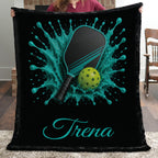 A person holds up a black fleece blanket featuring a custom pickleball design with a teal splash, a paddle, a yellow-green ball, and the name 'Trena' in teal script. The background shows a home interior with a wooden chair and rug.
