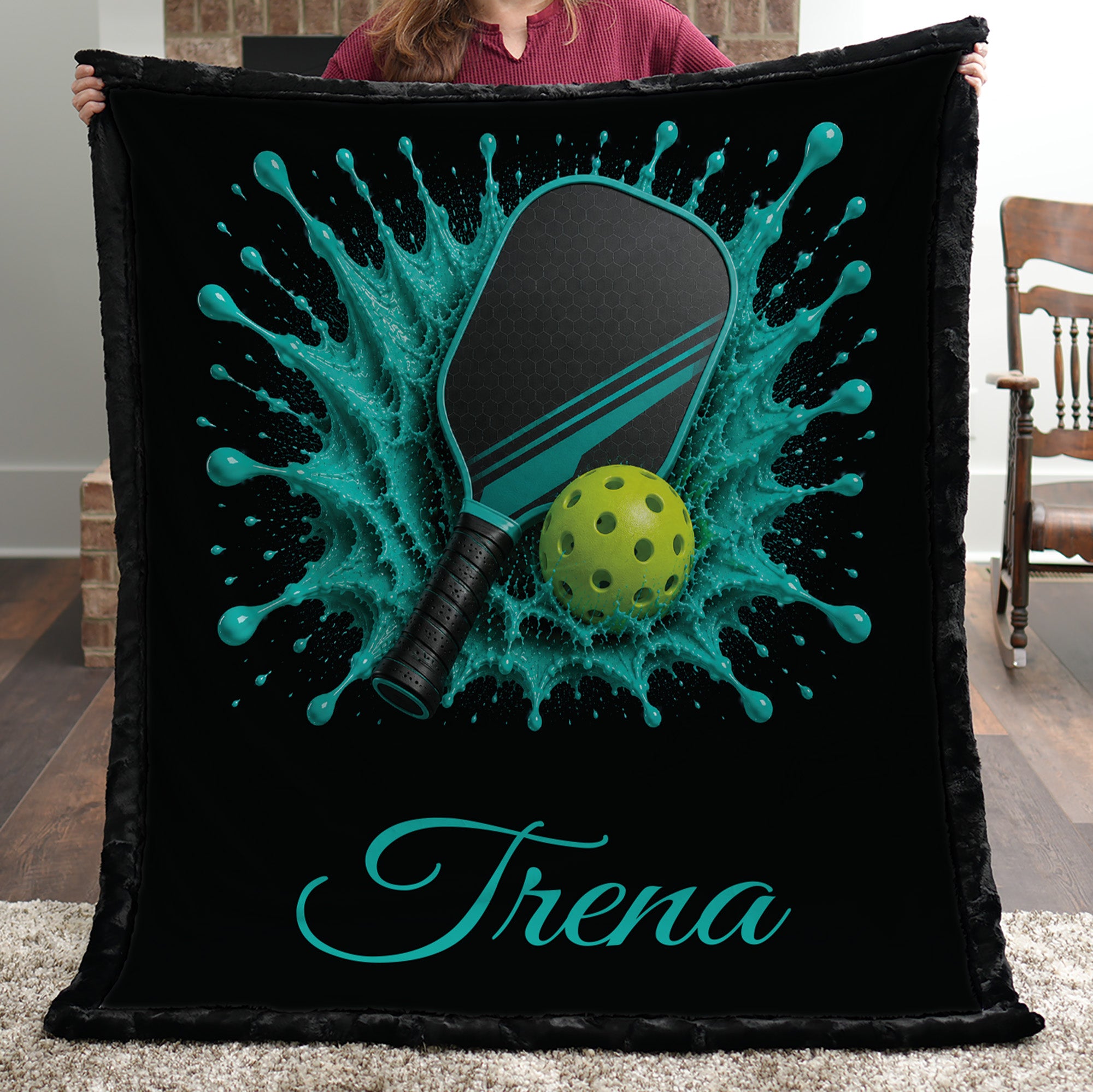 A person holds up a black fleece blanket featuring a custom pickleball design with a teal splash, a paddle, a yellow-green ball, and the name 'Trena' in teal script. The background shows a home interior with a wooden chair and rug.