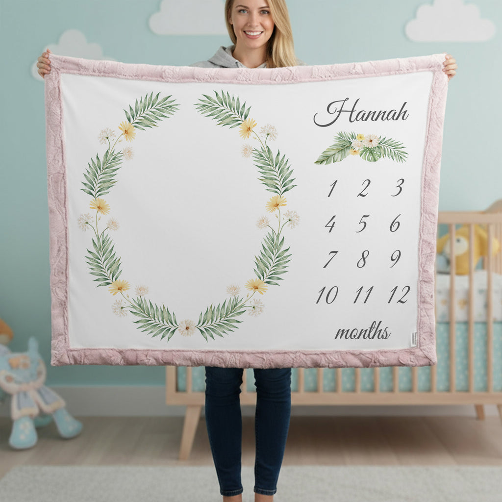 A woman holds up a personalized baby blanket featuring a pink border, a green leaf wreath with small yellow flowers, and a customizable name and monthly calendar grid on a white background. The setting is a nursery with a crib and light blue walls in the background.