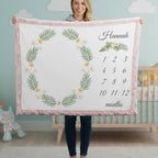 A woman holds up a personalized baby blanket featuring a pink border, a green leaf wreath with small yellow flowers, and a customizable name and monthly calendar grid on a white background. The setting is a nursery with a crib and light blue walls in the background.