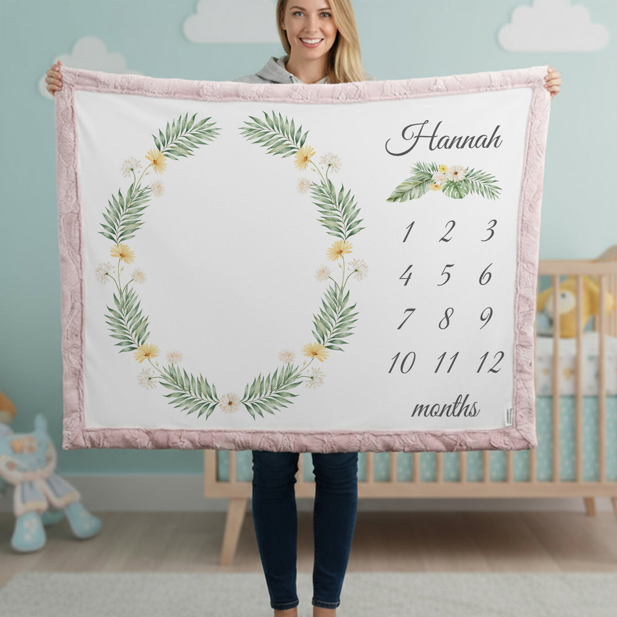 A woman holds up a personalized baby blanket featuring a pink border, a green leaf wreath with small yellow flowers, and a customizable name and monthly calendar grid on a white background. The setting is a nursery with a crib and light blue walls in the background.