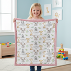 A young girl stands in a bright children's room holding up a square baby blanket. The blanket has a pink border and a white center printed with cute cartoon animals including elephants, giraffes, and monkeys, along with the name 'COOPER' repeated.