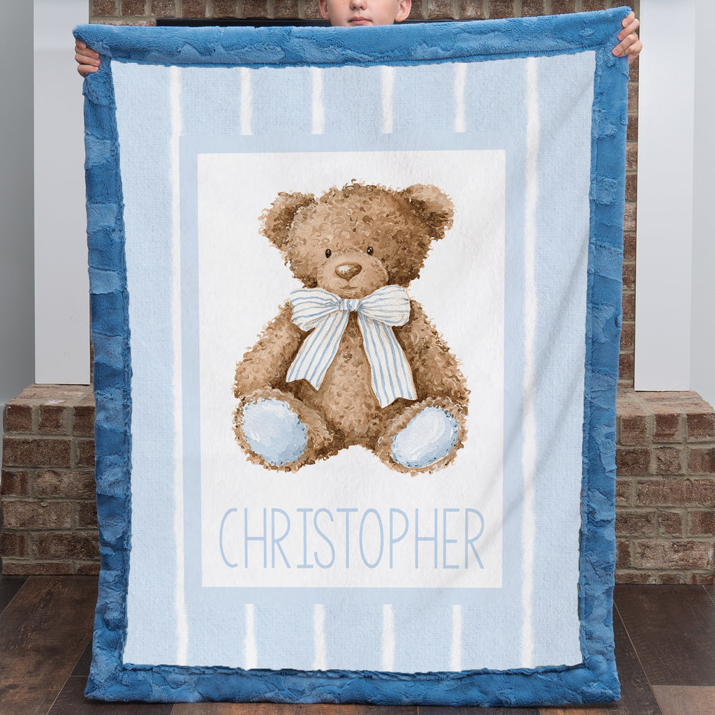 A baby blanket held up by a child, featuring a brown teddy bear with a blue and white striped bow on a light blue and white striped background, with the name 'CHRISTOPHER' printed at the bottom. The blanket has a textured blue border.