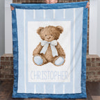 A baby blanket held up by a child, featuring a brown teddy bear with a blue and white striped bow on a light blue and white striped background, with the name 'CHRISTOPHER' printed at the bottom. The blanket has a textured blue border.