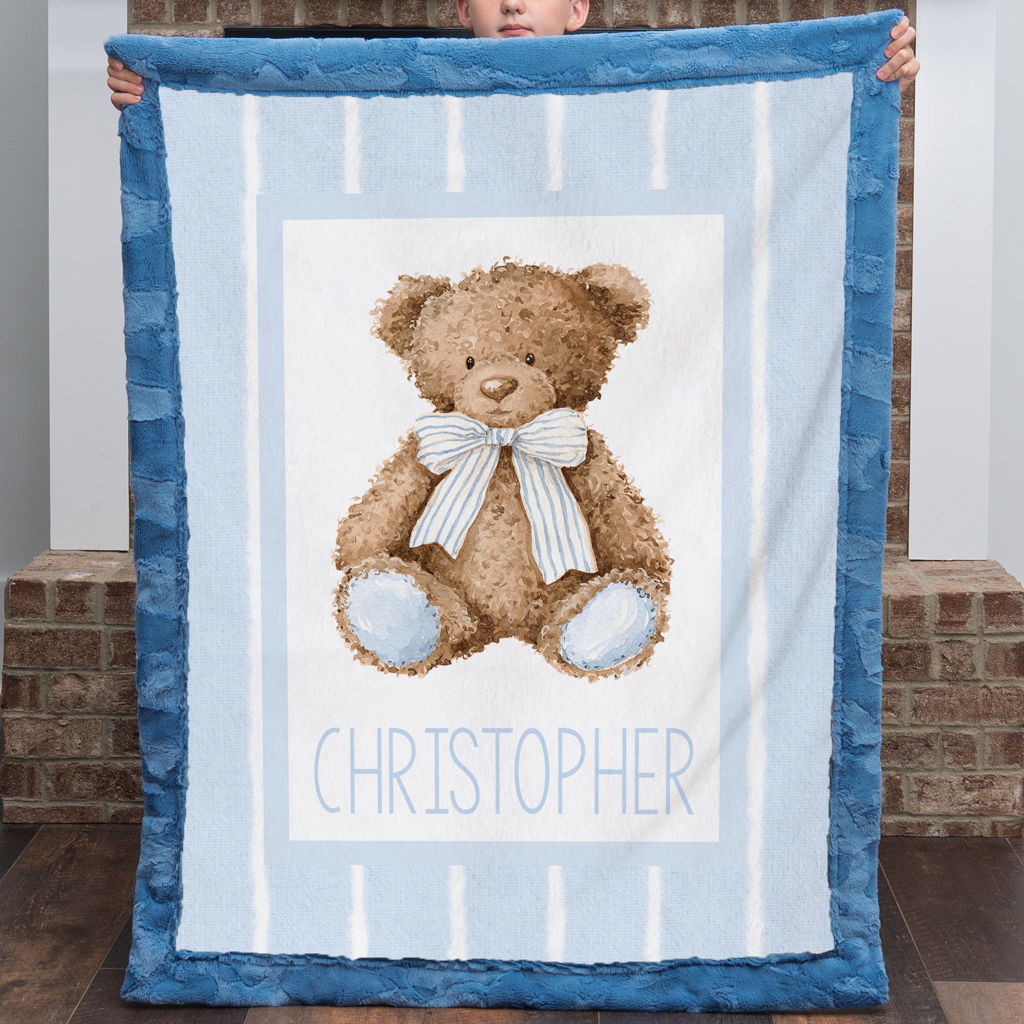 A baby blanket held up by a child, featuring a brown teddy bear with a blue and white striped bow on a light blue and white striped background, with the name 'CHRISTOPHER' printed at the bottom. The blanket has a textured blue border.