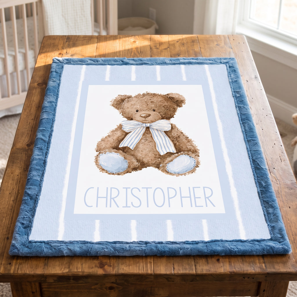 A personalized baby blanket featuring a watercolor teddy bear with a striped bow tie and the name 'CHRISTOPHER' printed below, displayed on a wooden table in a nursery setting.