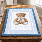 A personalized baby blanket featuring a watercolor teddy bear with a striped bow tie and the name 'CHRISTOPHER' printed below, displayed on a wooden table in a nursery setting.