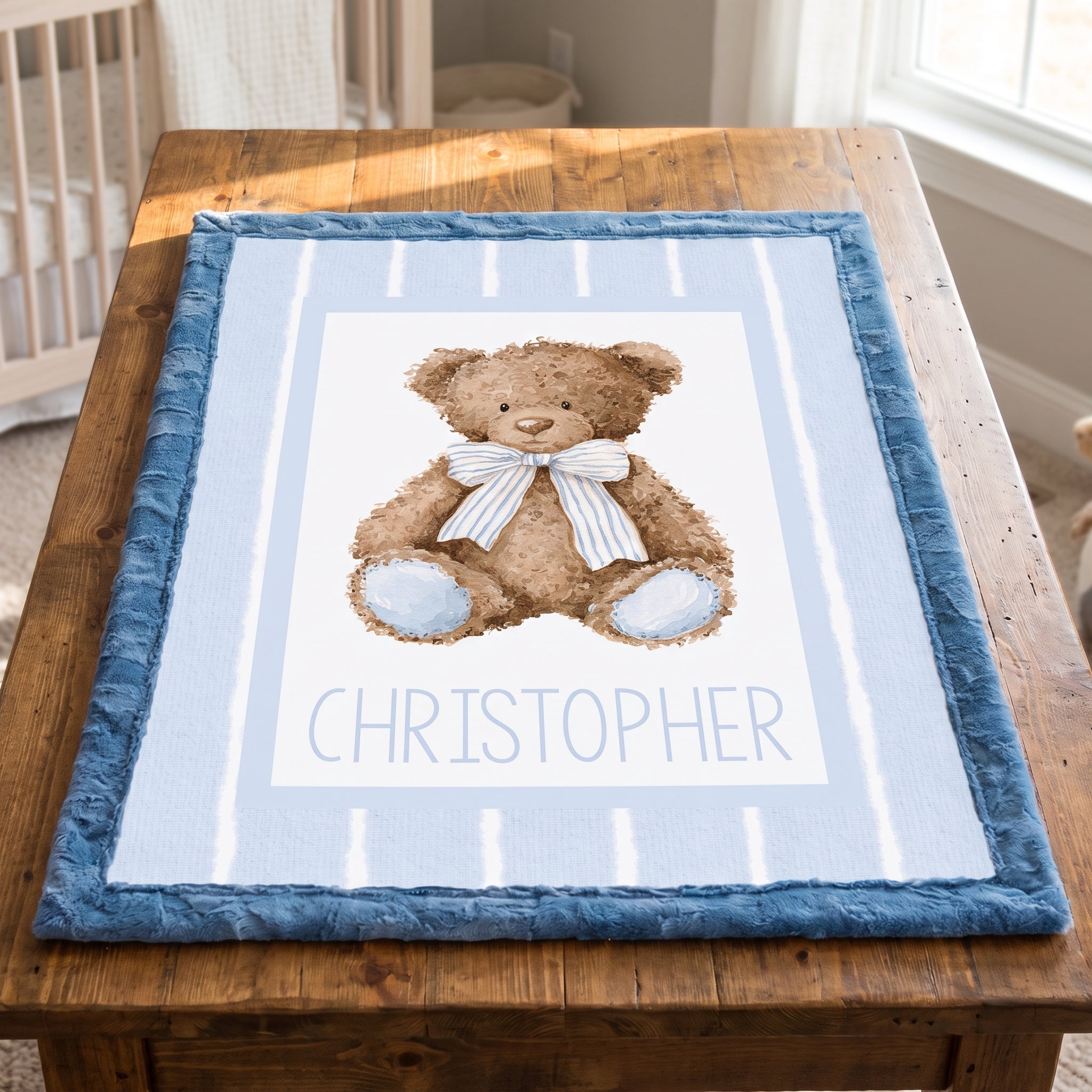 A personalized baby blanket featuring a watercolor teddy bear with a striped bow tie and the name 'CHRISTOPHER' printed below, displayed on a wooden table in a nursery setting.