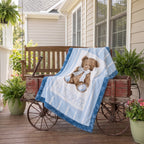 A personalized baby blanket draped over a rustic wooden cart on a front porch. The blanket features a light blue and white striped background, a brown teddy bear wearing a blue bow tie, and the name 'CHRISTOPHER' printed in blue.