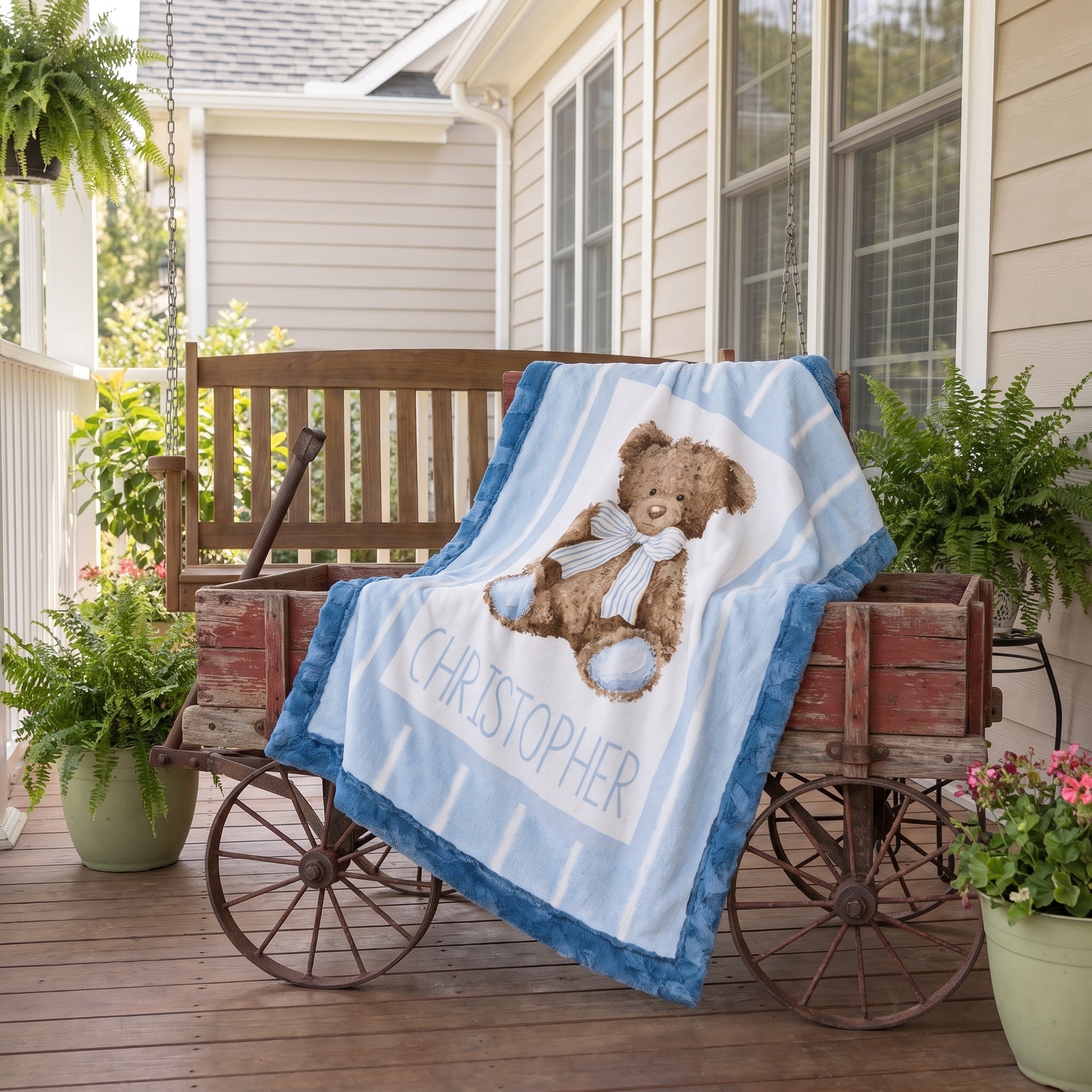 A personalized baby blanket draped over a rustic wooden cart on a front porch. The blanket features a light blue and white striped background, a brown teddy bear wearing a blue bow tie, and the name 'CHRISTOPHER' printed in blue.
