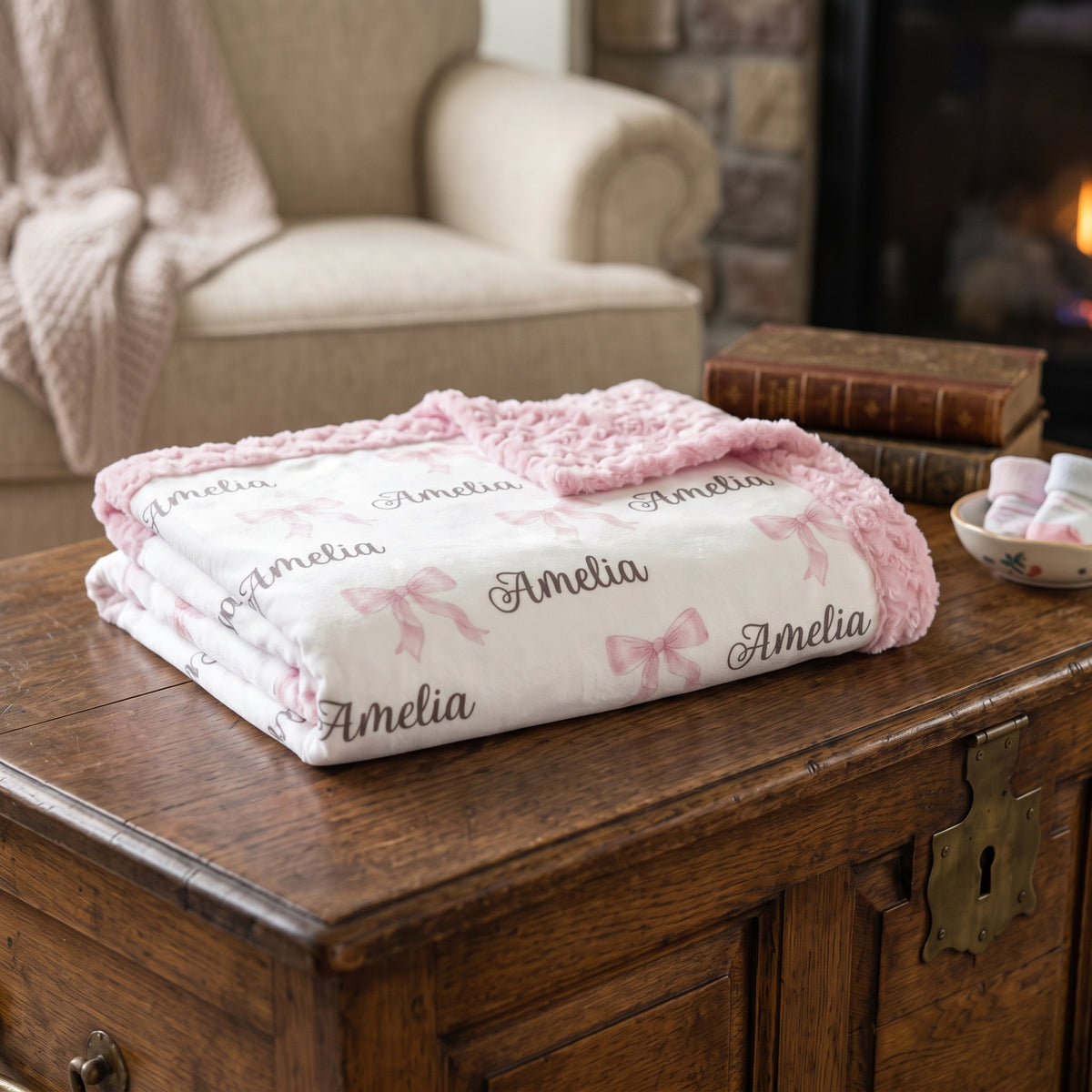 A folded white blanket with pink ruffle trim and pink bow motifs, printed with the name 'Amelia' in script, displayed on a rustic wooden chest in a cozy living room setting.