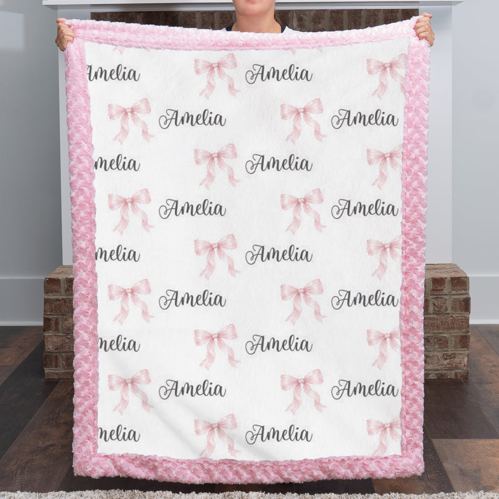 A person holds up a personalized fleece blanket with a pink fuzzy border. The blanket has a white center printed with pink bows and the name 'Amelia' in black script.