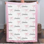 A person holds up a personalized fleece blanket with a pink fuzzy border. The blanket has a white center printed with pink bows and the name 'Amelia' in black script.