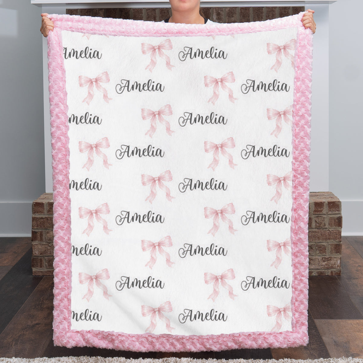 A person holds up a personalized fleece blanket with a pink fuzzy border. The blanket has a white center printed with pink bows and the name 'Amelia' in black script.