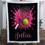 A personalized throw blanket with a white ruffled border is held up by a person. The blanket features a black background with a vibrant pink tennis splash, a black tennis racket, a yellow tennis ball, and the name 'Julia' in pink script.
