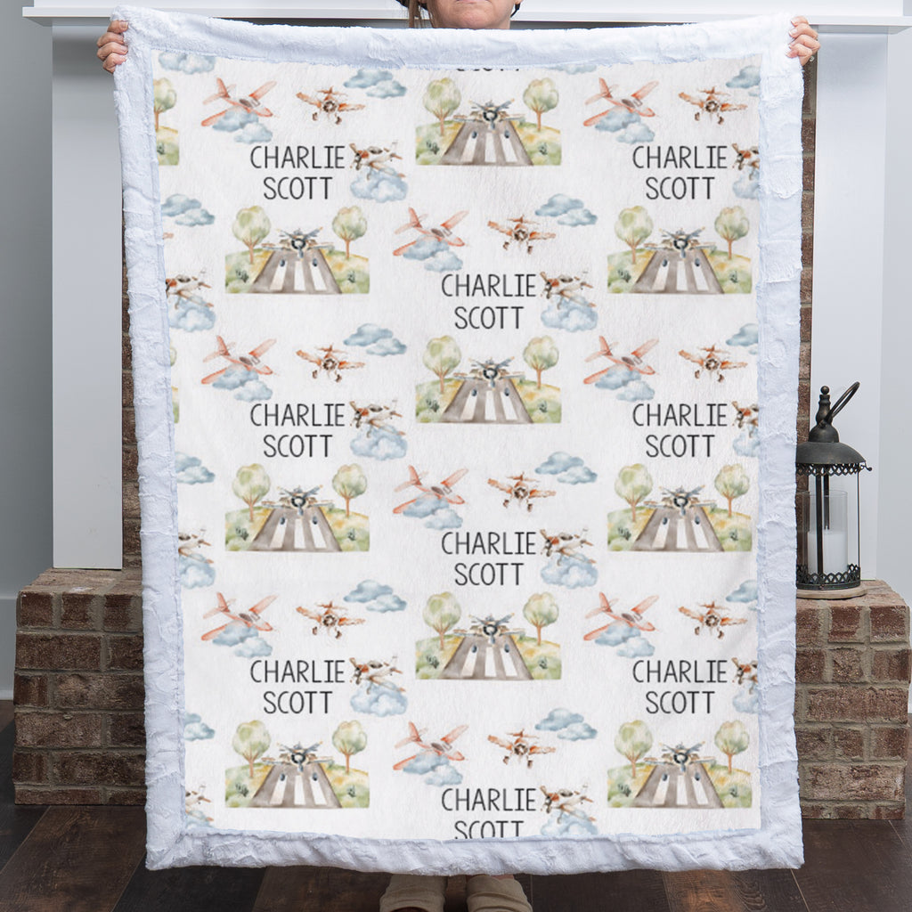 A person holds up a personalized quilted blanket featuring a repeating pattern of vintage airplanes, trees, and the name 'CHARLIE SCOTT' printed in black. The blanket has a light blue ruffled border and is shown against a home entryway background with a brick pillar and lantern.