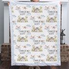 A person holds up a personalized quilted blanket featuring a repeating pattern of vintage airplanes, trees, and the name 'CHARLIE SCOTT' printed in black. The blanket has a light blue ruffled border and is shown against a home entryway background with a brick pillar and lantern.