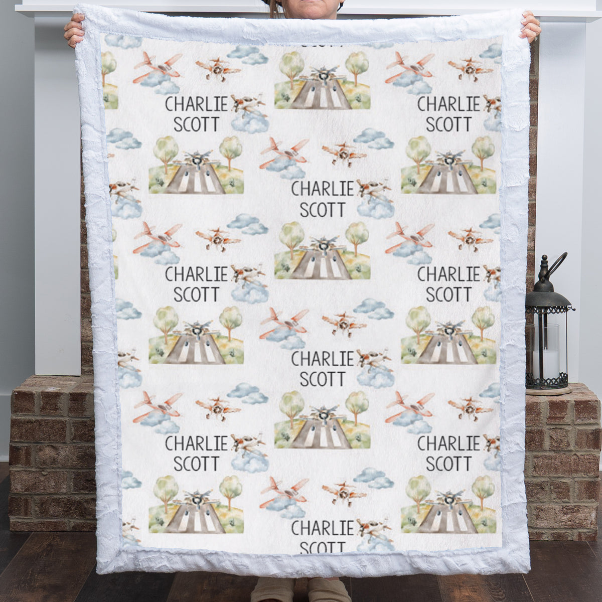 A person holds up a personalized quilted blanket featuring a repeating pattern of vintage airplanes, trees, and the name 'CHARLIE SCOTT' printed in black. The blanket has a light blue ruffled border and is shown against a home entryway background with a brick pillar and lantern.