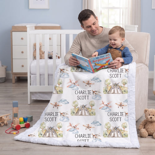 A father and child sitting on a beige armchair in a nursery, reading a book together while wrapped in a white plush blanket printed with personalized names and airplane illustrations. A white crib, wooden dresser, toys, and teddy bears are visible in the room.