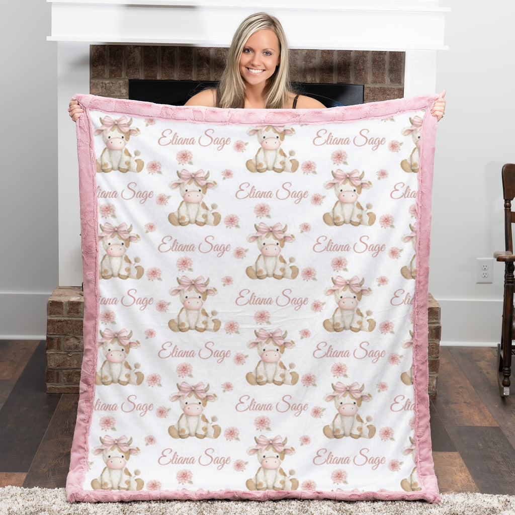 Personalized highland cow minky baby blanket with bright pink fur trim, held up by fireplace