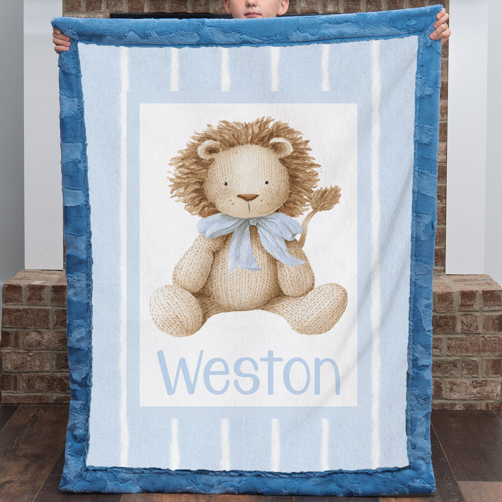 A person holds up a personalized plush blanket featuring a teddy bear design and the name 'Weston' printed at the bottom. The blanket has a blue border and a light blue and white striped center with a beige teddy bear wearing a blue bow tie.