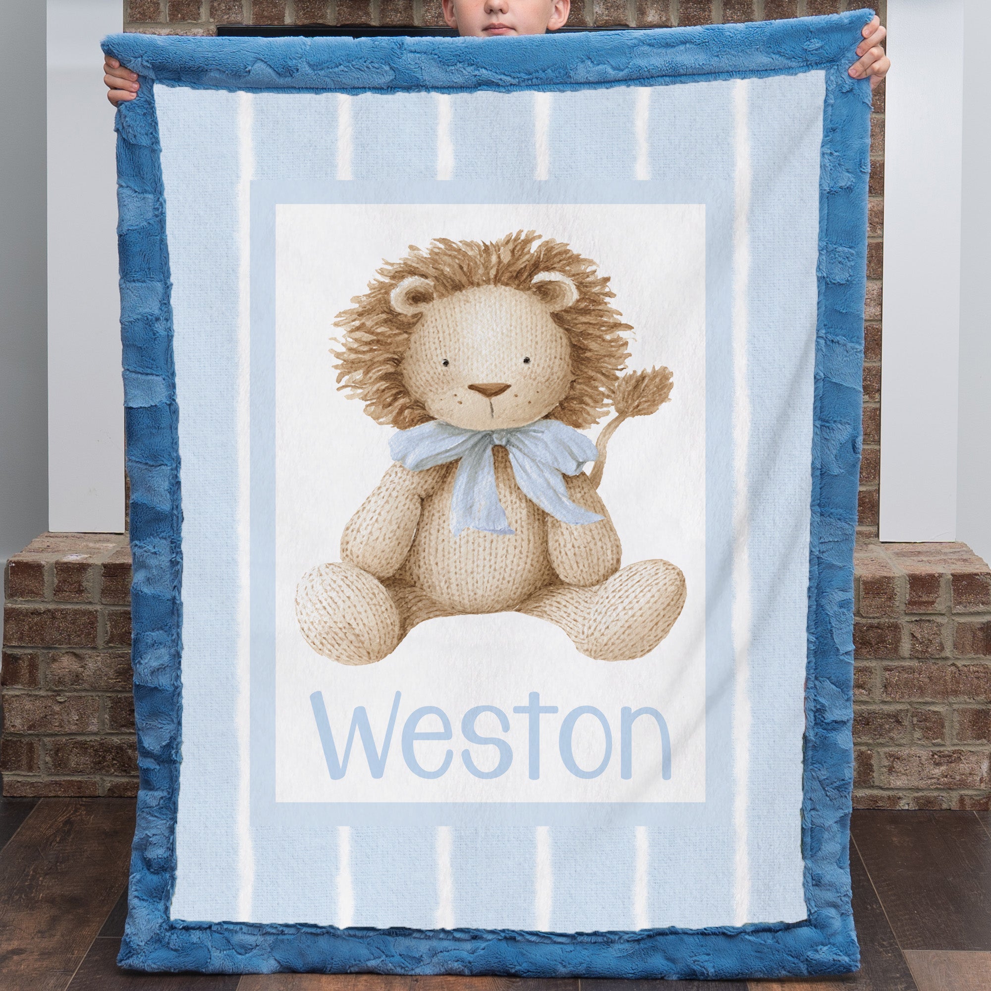 A person holds up a personalized plush blanket featuring a teddy bear design and the name 'Weston' printed at the bottom. The blanket has a blue border and a light blue and white striped center with a beige teddy bear wearing a blue bow tie.