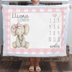 A person holds up a personalized baby blanket featuring a pink gingham border, a knitted elephant illustration with a pink bow, the names 'Eliana' and 'Elizabeth', and a pink numbered months calendar from 1 to 12.