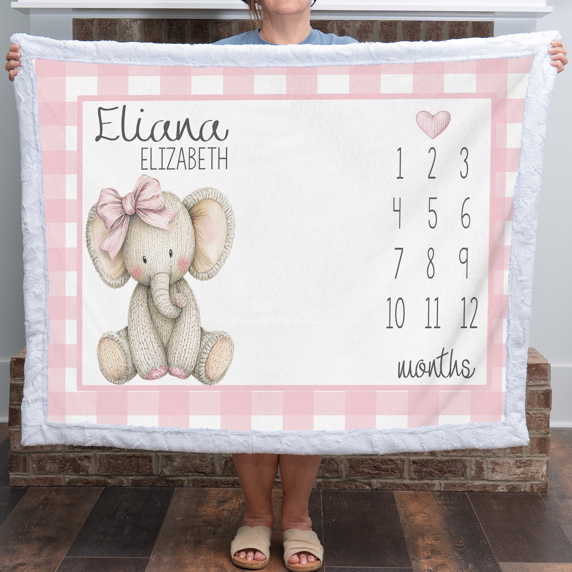 A person holds up a personalized baby blanket featuring a pink gingham border, a knitted elephant illustration with a pink bow, the names 'Eliana' and 'Elizabeth', and a pink numbered months calendar from 1 to 12.