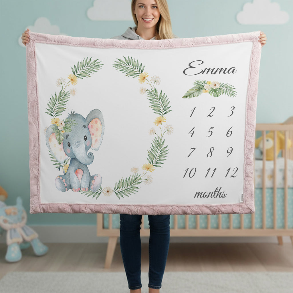 A woman holds up a personalized baby blanket in a nursery setting. The blanket features a pink border, a watercolor gray elephant with a floral wreath, a floral design with the name 'Emma', and a 12-month calendar grid.