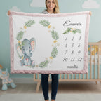 A woman holds up a personalized baby blanket in a nursery setting. The blanket features a pink border, a watercolor gray elephant with a floral wreath, a floral design with the name 'Emma', and a 12-month calendar grid.