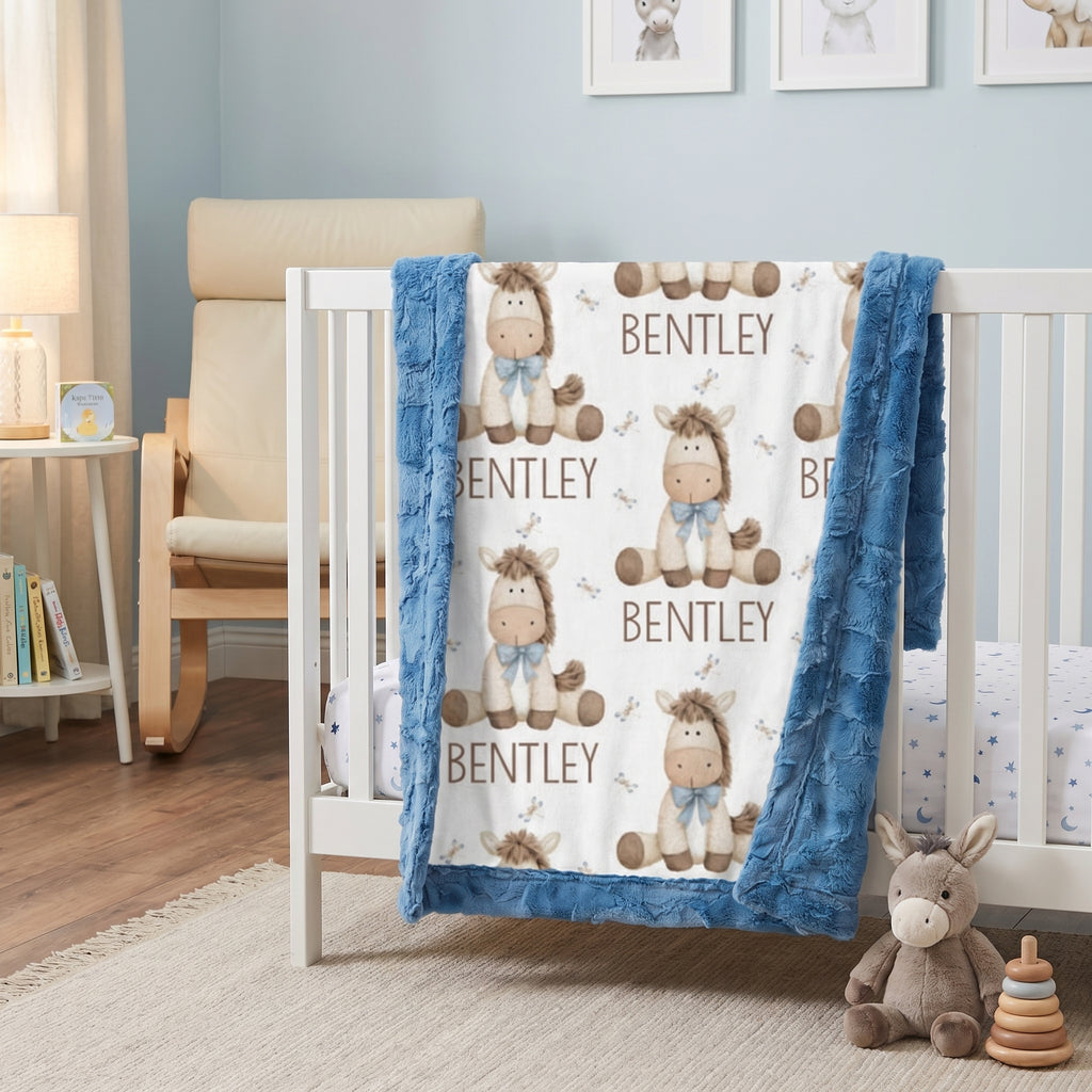 A personalized baby blanket with a blue faux-fur border and a white center printed with cartoon donkeys and the name 'BENTLEY', draped over a white crib in a styled nursery setting.