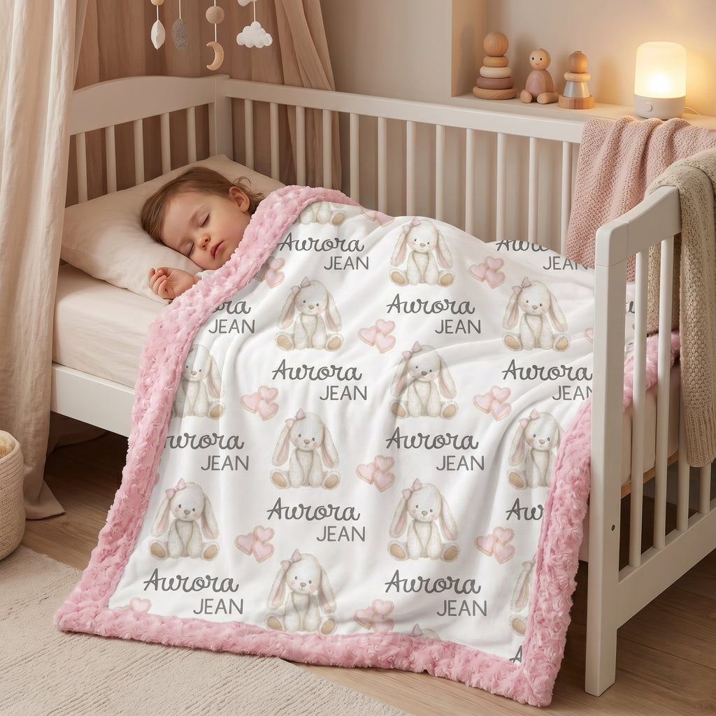 A baby sleeping in a white wooden crib, covered with a white blanket featuring a repeating pattern of gray bunny illustrations, the text 'Aurora JEAN', and a soft pink fuzzy border.