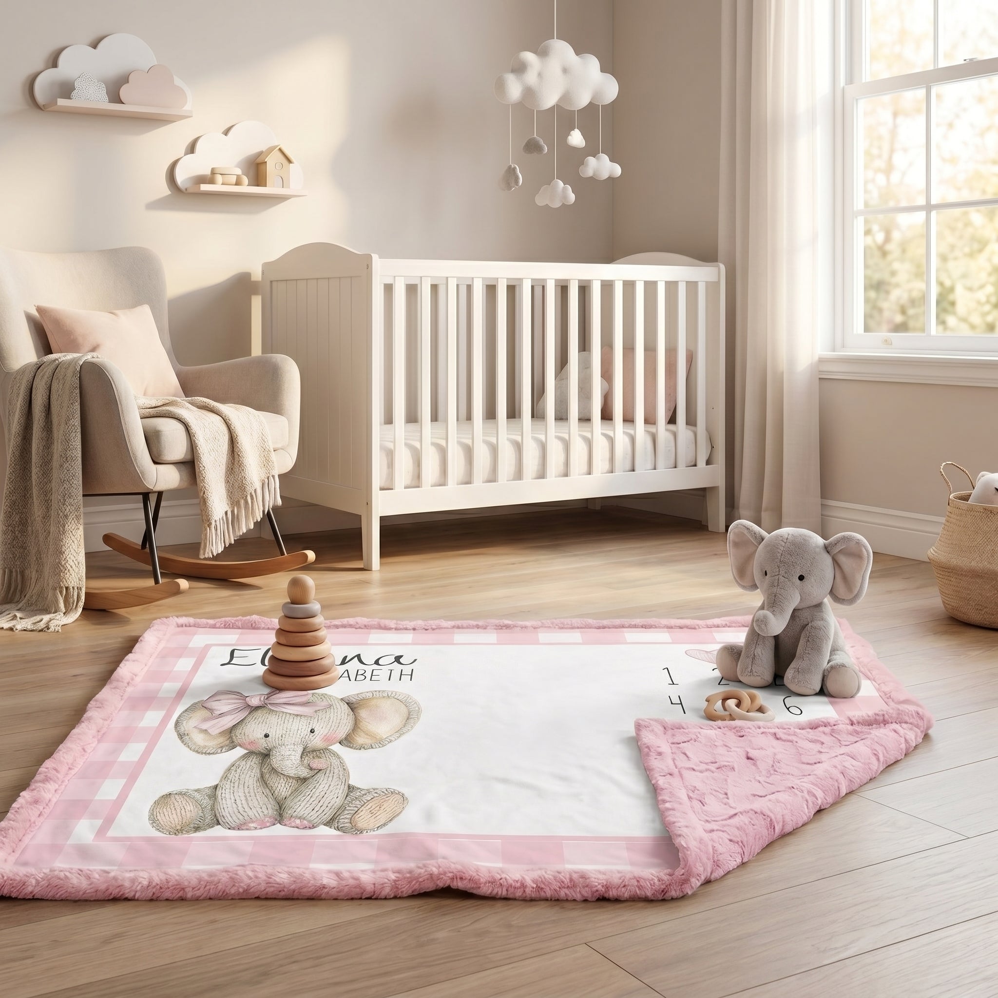 A pink and white baby quilt with a plush elephant design and the text 'BONSAI ARETH' is spread out on a wooden floor in a styled nursery. A gray plush elephant toy sits on the corner of the quilt, with a white crib, rocking chair, and window in the background.