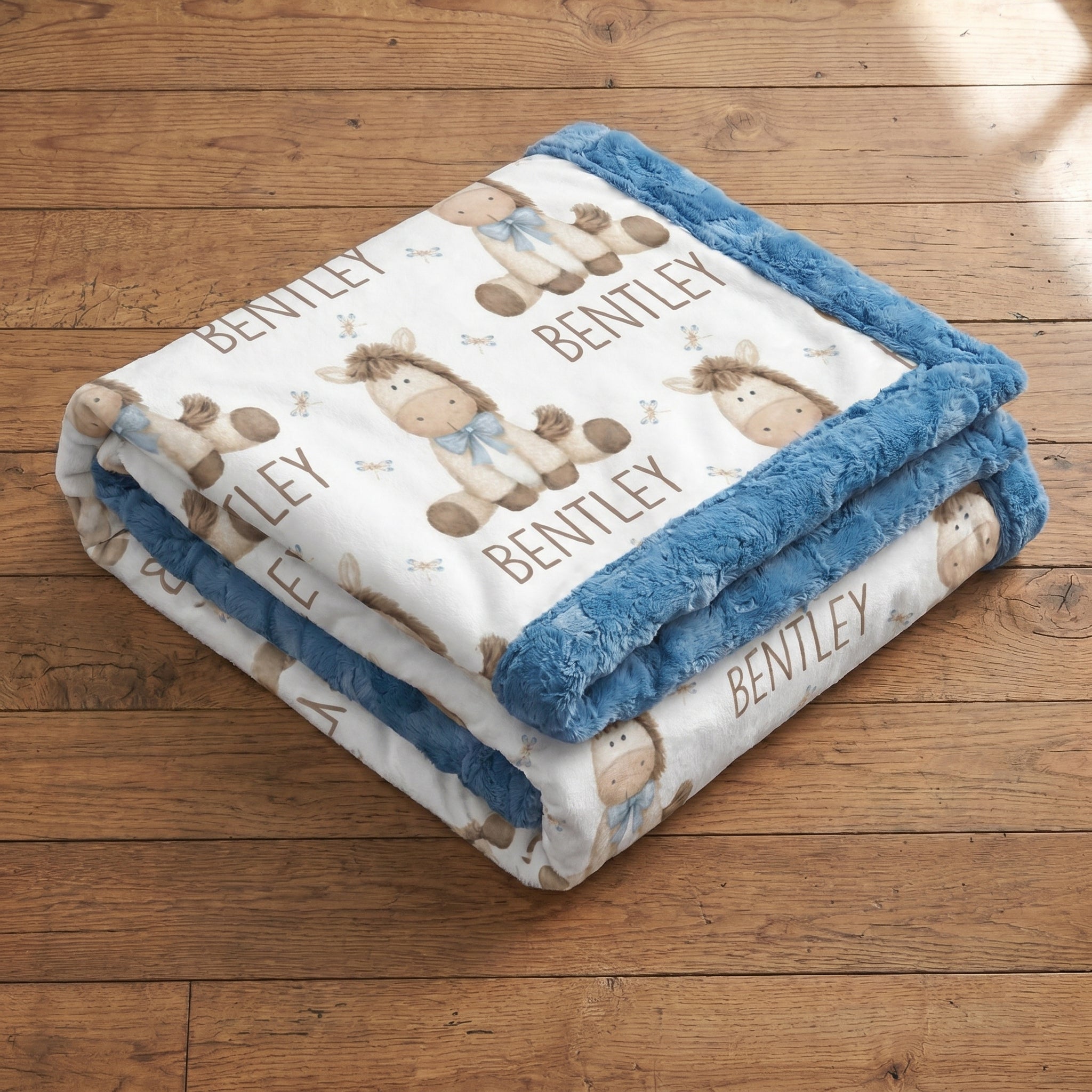 A folded baby blanket with a blue plush border and a white center printed with brown horse illustrations and the name 'BENTLEY' in tan lettering, displayed on a wooden floor.