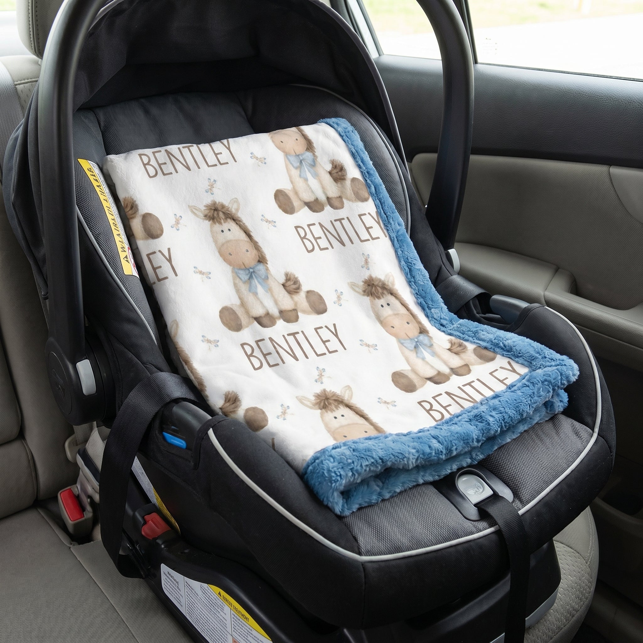 A personalized baby car seat blanket with a teddy bear pattern and the name 'BENTLEY' printed repeatedly, laid out inside a black infant car seat on a car seat.