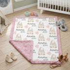 A personalized baby blanket with a pink border, featuring a white center printed with bunny illustrations, pink hearts, and the name 'Aurora JEAN' repeated. The blanket is styled in a nursery setting on a light wood floor with baby shoes, a stuffed elephant toy, and a crib in the background.