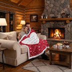A couple sits on a beige sofa in a cozy log-cabin living room, wrapped in a red and white fleece blanket with white text reading 'I Love You Through Love & Life' and small heart motifs. A stone fireplace with a burning fire is in the background, along with a coffee table and warm ambient lighting.