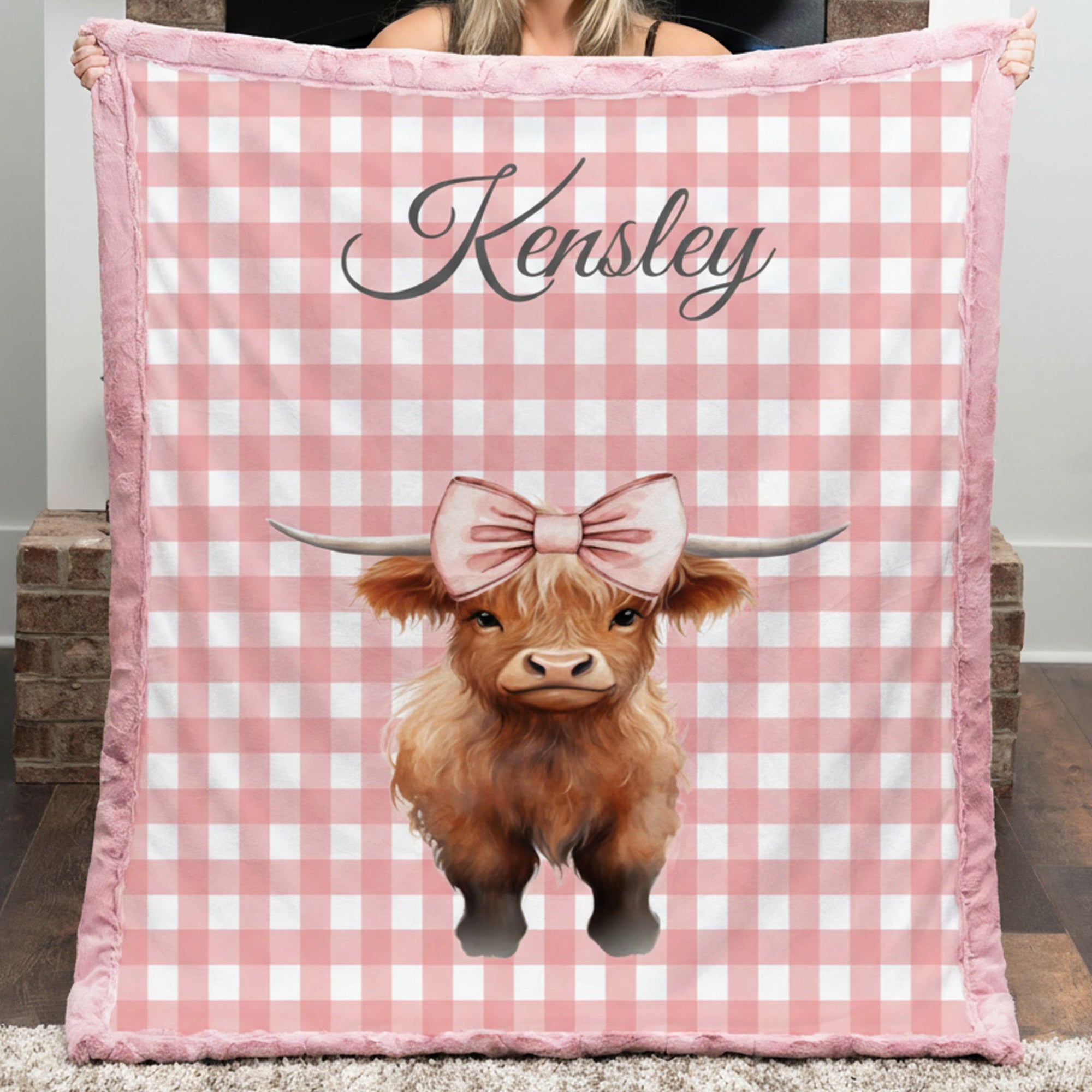 Person holding a pink checkered blanket with a cow illustration and 'Kensley' text.