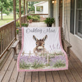 A personalized blanket draped over a wooden porch swing on a rustic wooden porch. The blanket features a white background with a printed illustration of a donkey behind a wooden fence in a field of purple lavender.